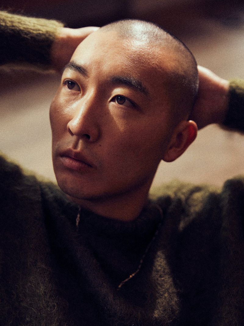 East Asian man with a buzzcut resting his hands behind his head while wearing an olive green fuzzy knit sweater.