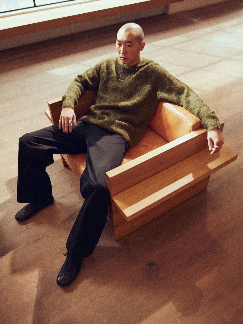 An East Asian man with a buzz cut wearing an olive green knit sweater seated in a wooden armchair.