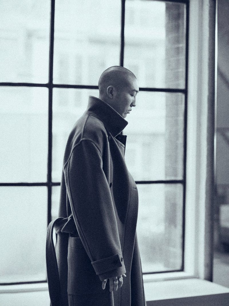 Man with a shaved head wearing an oversized dark wool coat standing in profile in front of large multi-pane grid windows.