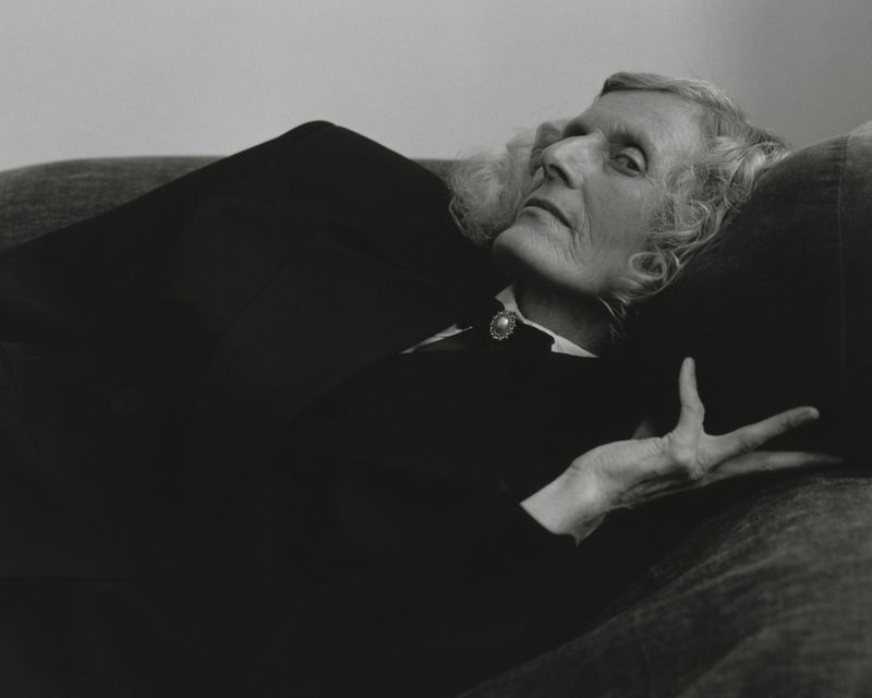 Elderly woman with wavy grey hair reclining on a dark sofa wearing a black blazer and collared shirt with a round brooch.
