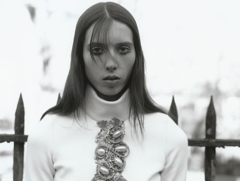Black and white portrait of a woman in a white turtleneck sweater with beaded jewel chest detailing in front of a metal fence.