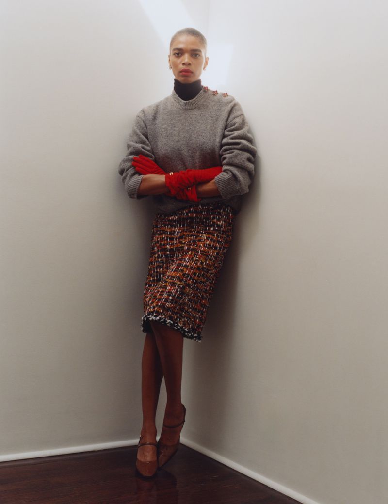 Model with short buzzed hair standing in a corner wearing a grey crewneck sweater, red gloves, and a patterned tweed skirt.
