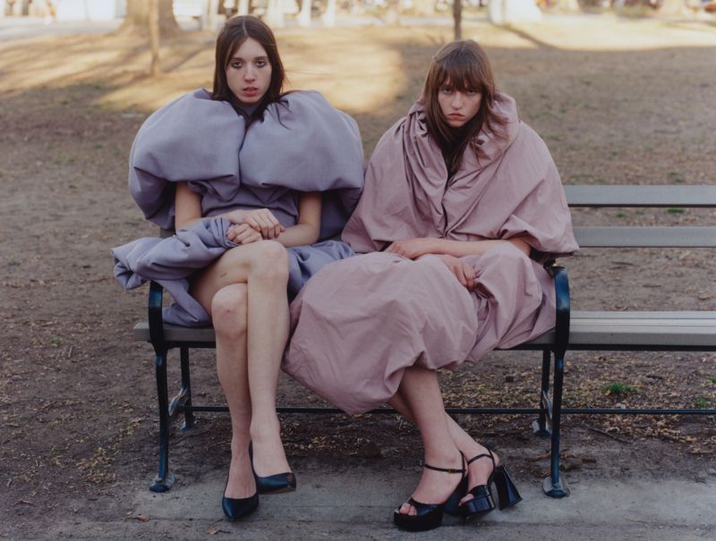 Two fashion models seated on an outdoor park bench wearing voluminous sculptural lavender and dusty rose dresses with black heels.