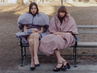 Two fashion models seated on an outdoor park bench wearing voluminous sculptural lavender and dusty rose dresses with black heels.