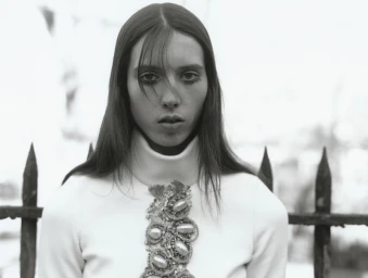 Black and white portrait of a woman in a white turtleneck sweater with beaded jewel chest detailing in front of a metal fence.
