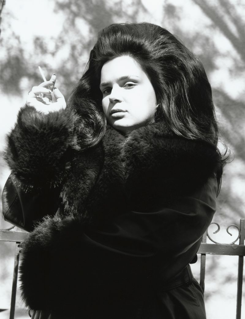 Monochrome portrait of a woman with voluminous 1960s bouffant hair holding a cigarette and wearing a dark fur-trimmed coat.