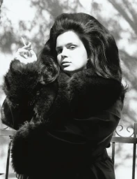Monochrome portrait of a woman with voluminous 1960s bouffant hair holding a cigarette and wearing a dark fur-trimmed coat.
