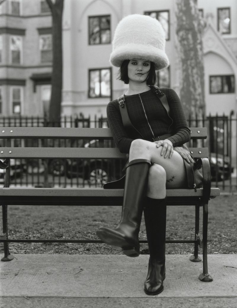 Model wearing an oversized white fuzzy dome hat and black leather boots seated on an outdoor wooden bench.