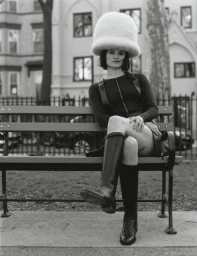Model wearing an oversized white fuzzy dome hat and black leather boots seated on an outdoor wooden bench.