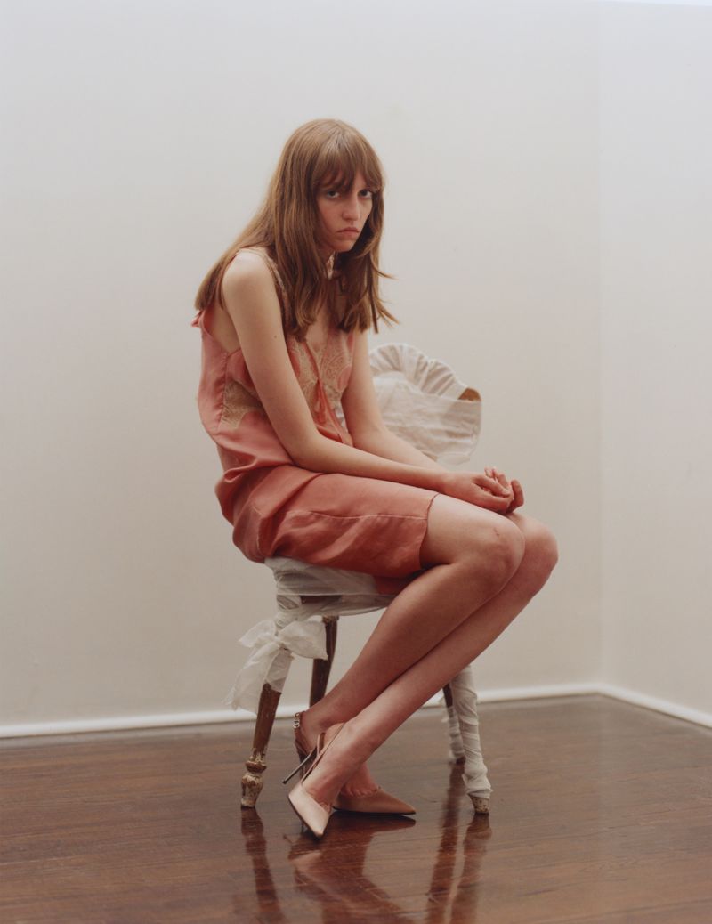 Woman in a pink slip dress seated on a fabric-wrapped wooden chair on polished hardwood floors against white walls.