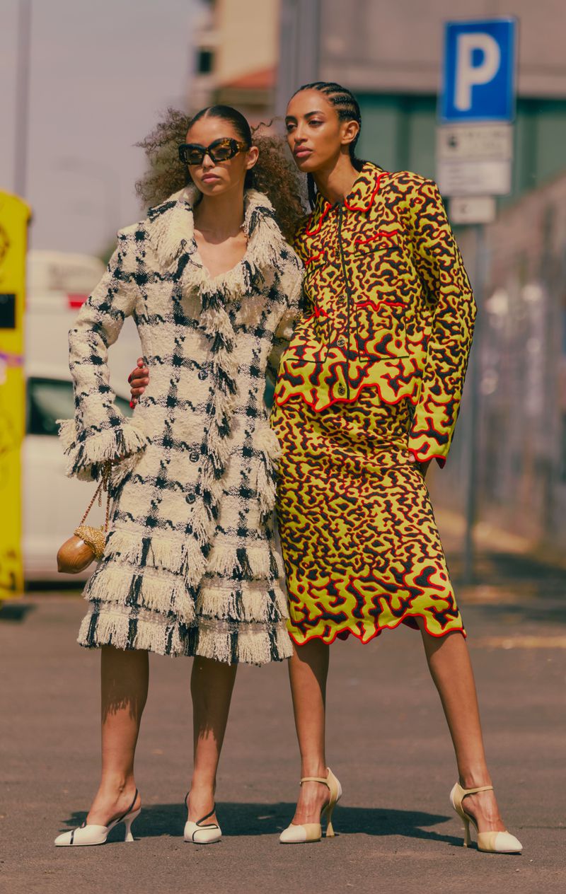 Two fashion models posing outdoors, one wearing a fringed plaid coat and sunglasses, the other in a vibrant yellow patterned skirt suit.