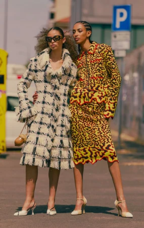 Two fashion models posing outdoors, one wearing a fringed plaid coat and sunglasses, the other in a vibrant yellow patterned skirt suit.