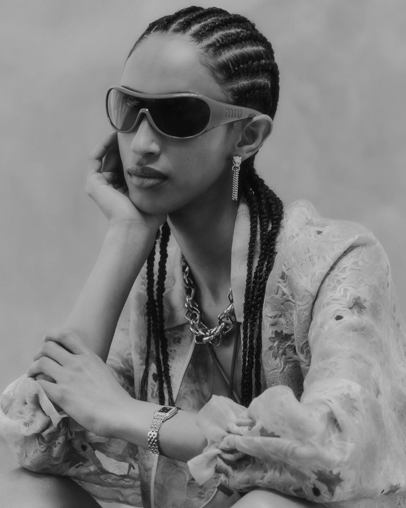 Model Yelibe Tilahun poses in dark wrap sunglasses, a sheer floral blouse, and jewellery against a grey studio backdrop.