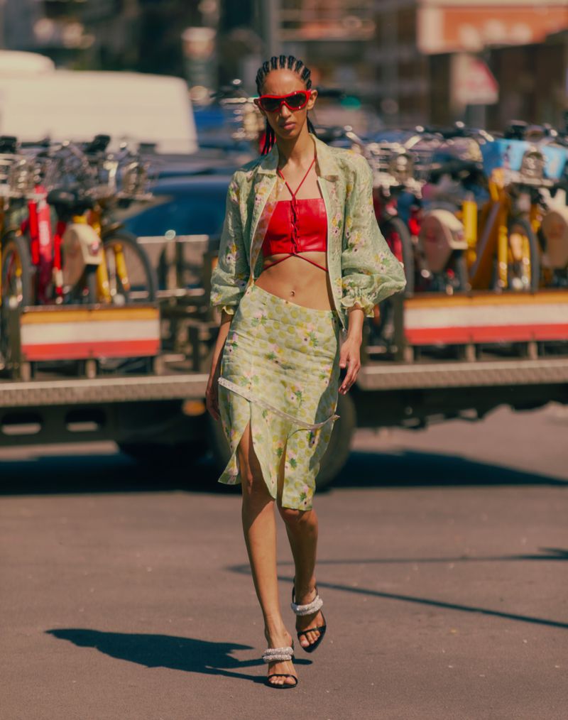 Model in green floral jacket and red strapless top walking on an urban street with parked bicycles in background.