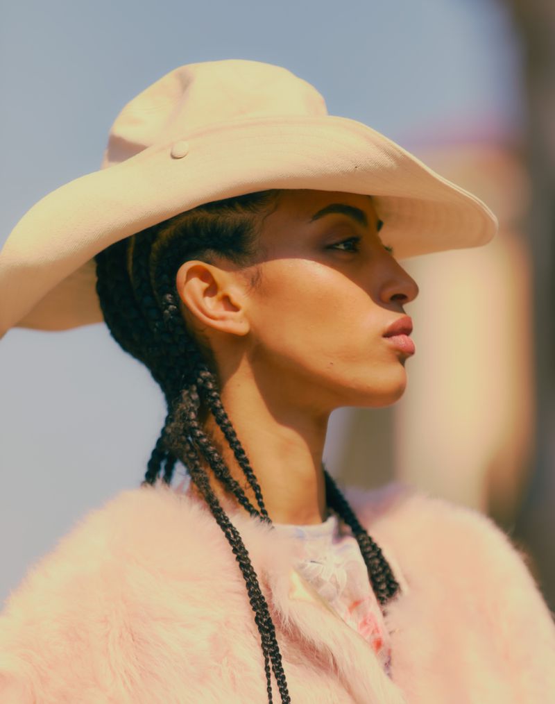 Model wearing a wide-brimmed cream hat and a fluffy pink jacket, seen in profile against an outdoor backdrop.