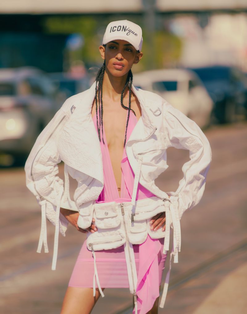 Model Yelibe Tilahun posing outdoors wearing a white cropped jacket, pink dress, and a white baseball cap reading ICONique.