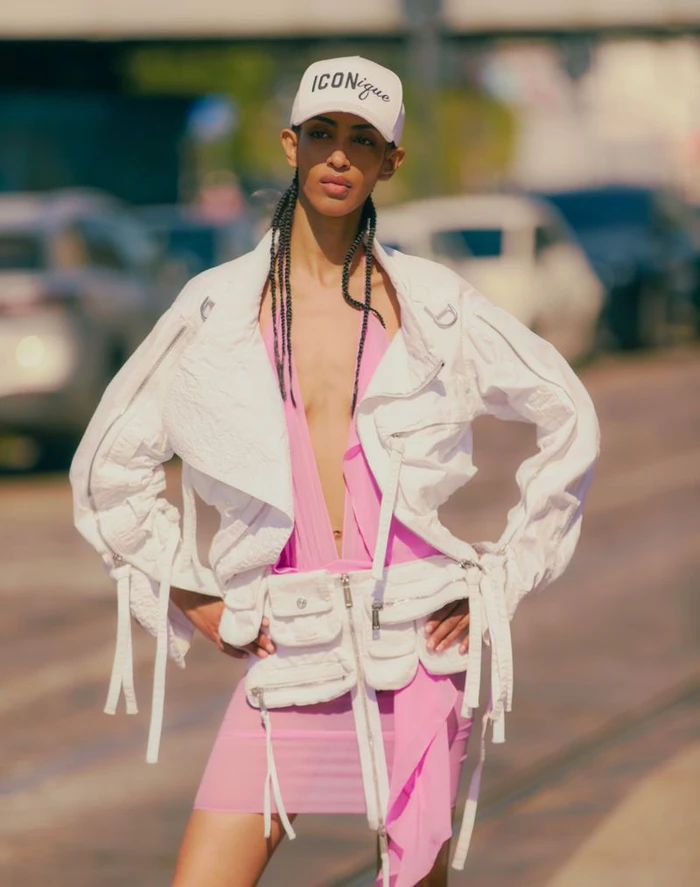 Model Yelibe Tilahun posing outdoors wearing a white cropped jacket, pink dress, and a white baseball cap reading ICONique.