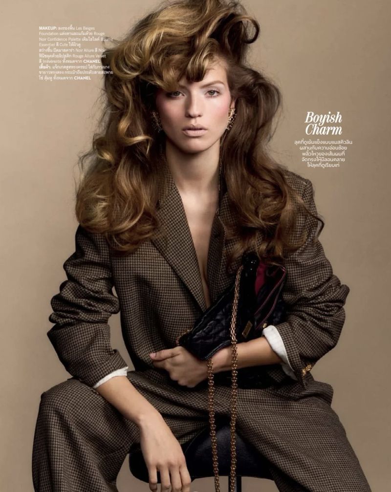 Model Carolina Mendillo wearing an oversized brown houndstooth suit while holding a quilted bag against a beige background.