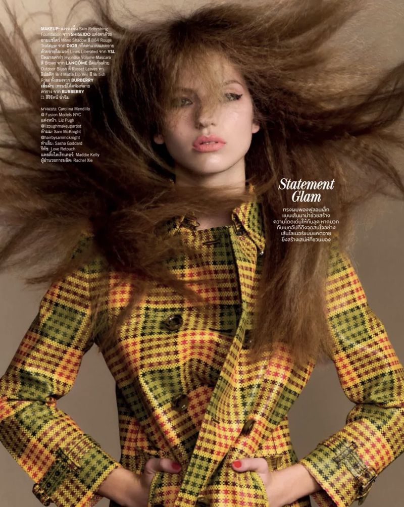 Model Carolina Mendillo with voluminous windswept hair posing in a yellow and olive plaid trench coat against a beige studio backdrop.