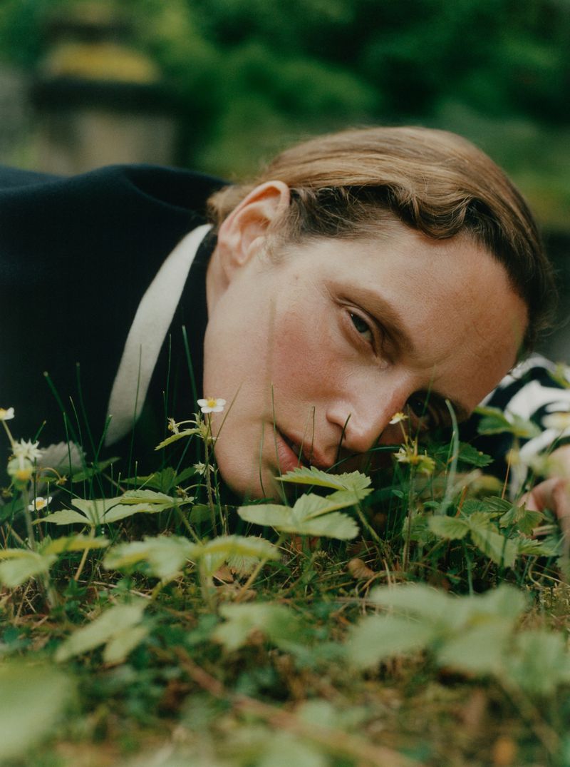 Close-up portrait of adult woman lying in grassy field with white flowers, wearing black jacket with white trim, in natural light.