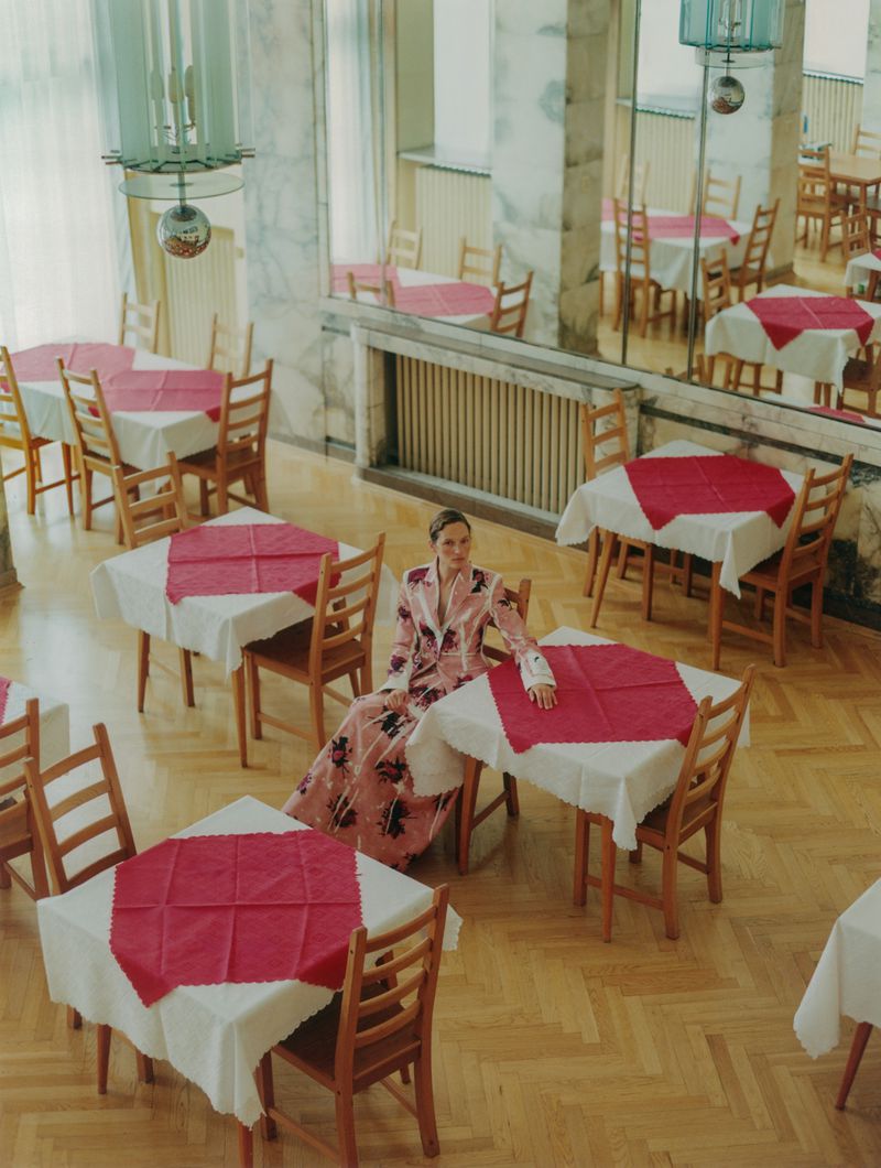 Overhead shot of a woman in a pink floral suit sitting at a table in an empty dining hall with red-and-white tablecloths and parquet floors.