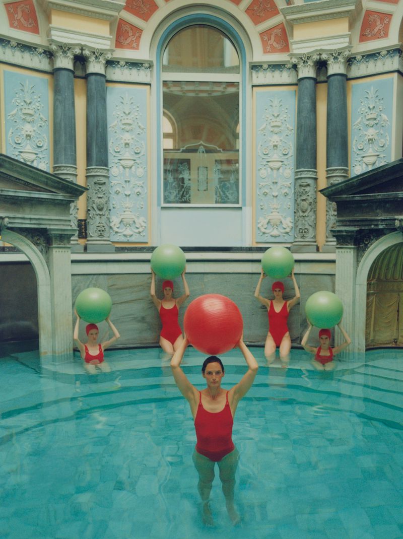 Group of women in red swimwear stand in a swimming pool, holding large exercise balls above their heads in a vintage interior.