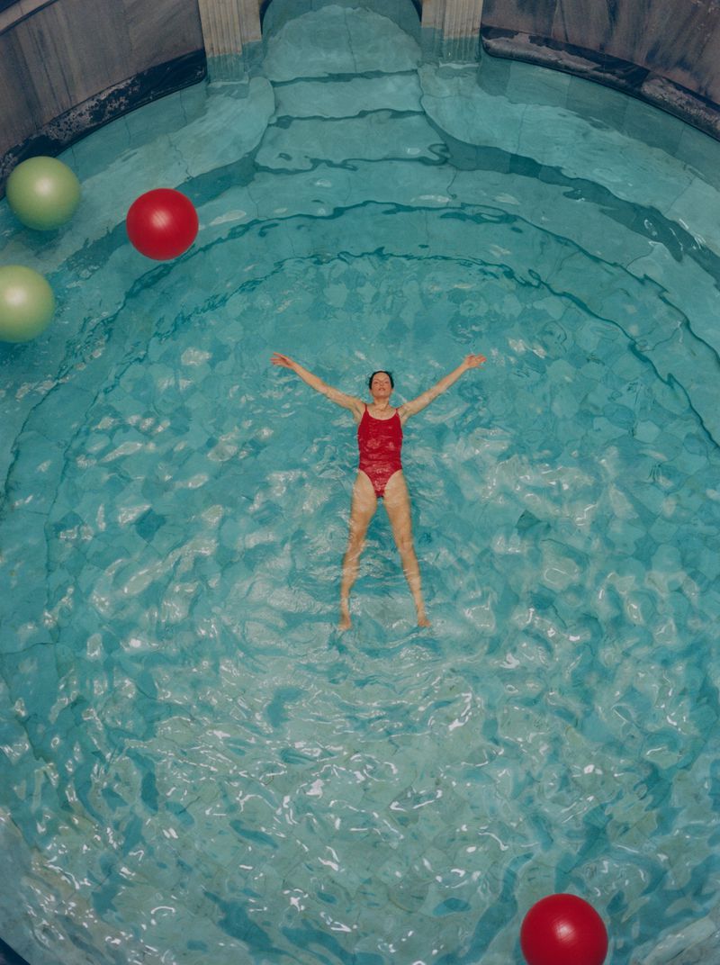 Woman in a red swimsuit floats in a pool surrounded by green and red spheres, with stone structures visible in the background.