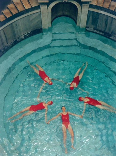 High-angle shot of five women in red swimsuits holding hands in a blue pool, creating a star formation, with stone architecture visible.