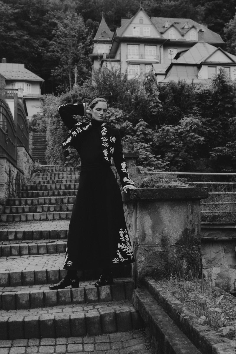 Woman in black embroidered dress on stone steps with stone railing, backed by trees and a house on a hill in black and white.