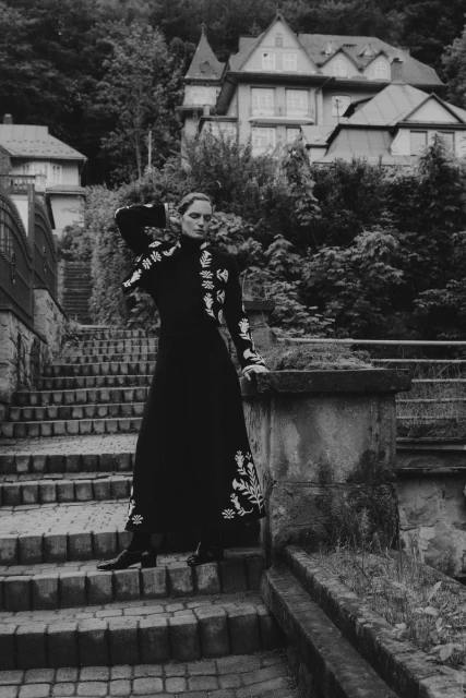 Woman in black embroidered dress on stone steps with stone railing, backed by trees and a house on a hill in black and white.