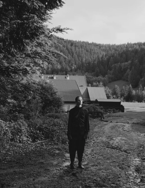 Monochrome portrait of a man standing in a rural setting with trees and a building in the background, captured in natural lighting.