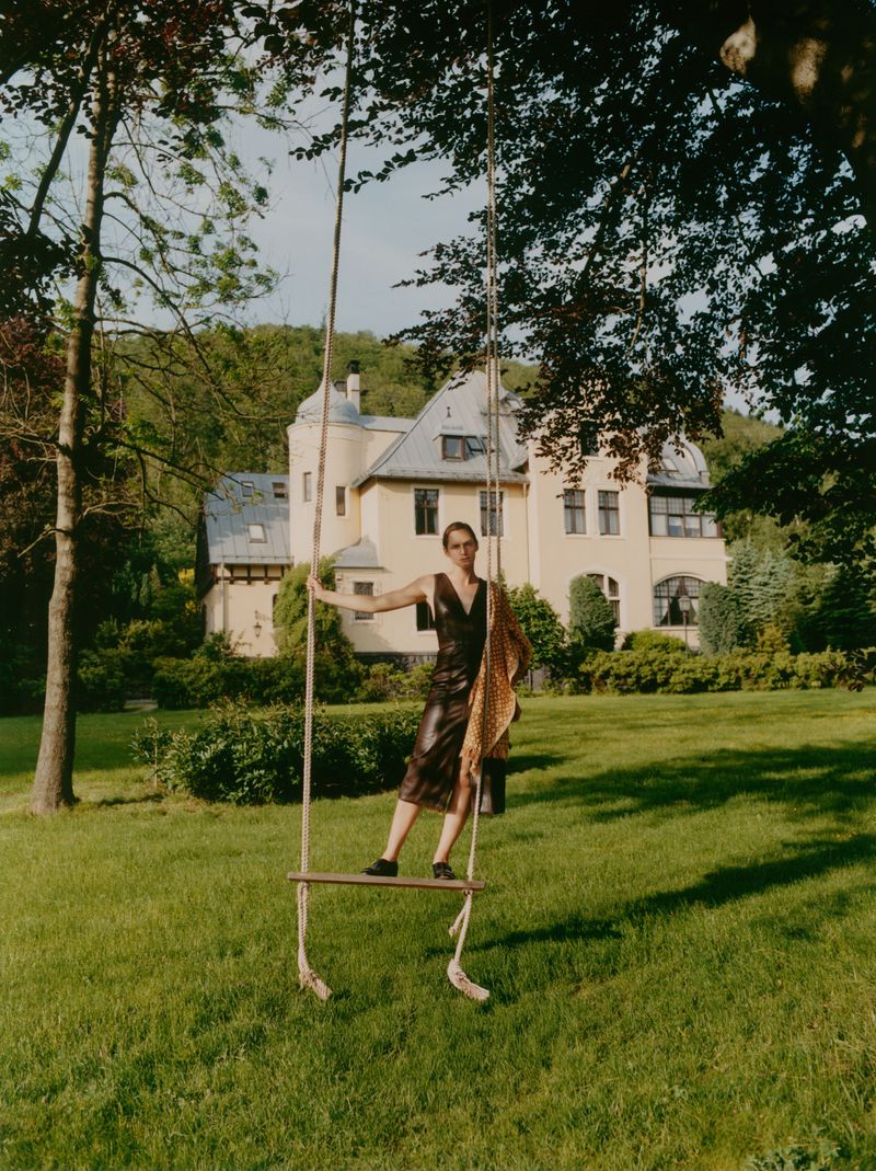 Woman in a dress stands on a rope swing in a grassy yard, with a large house, trees, and greenery in the background on a sunny day.