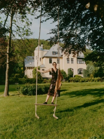 Woman in a dress stands on a rope swing in a grassy yard, with a large house, trees, and greenery in the background on a sunny day.