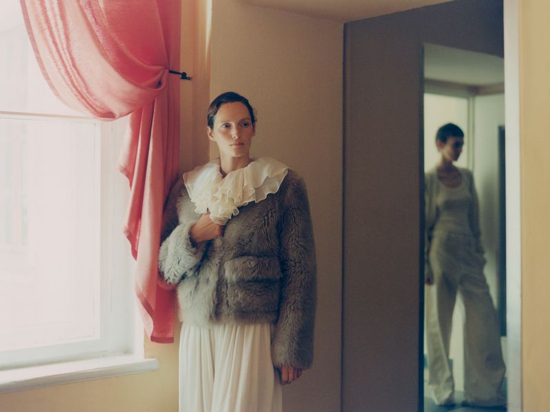 A woman in a fur jacket and white dress stands in a room, looking toward a window with pink curtains; another woman visible in doorway.