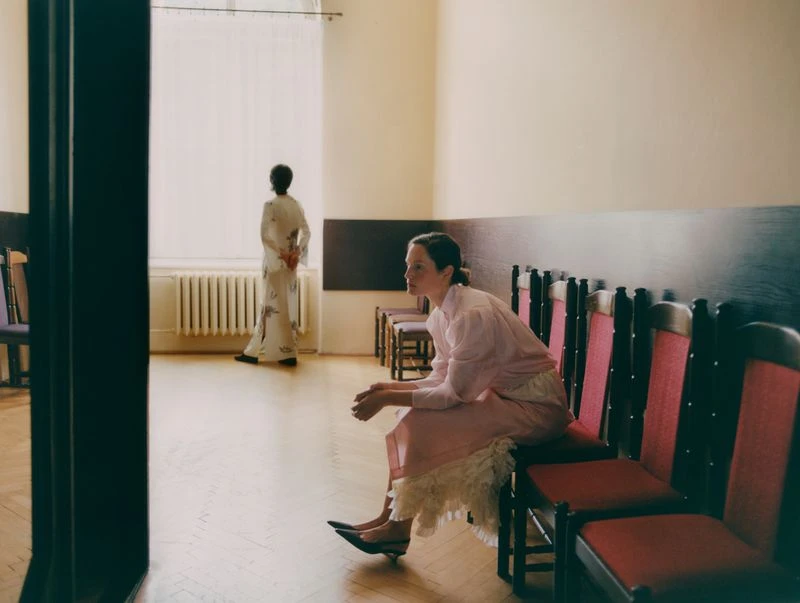 Two women in a sparse indoor setting, one sitting in foreground with a light pink dress, another standing near a window, wearing a floral robe.