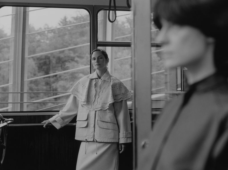 Monochrome shot of two women riding a tram; one stands at a window wearing a jacket, the other is blurred in the foreground.