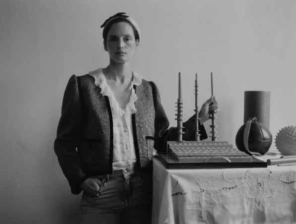 Monochrome portrait of a woman in a patterned jacket standing indoors, holding an architectural model, next to various cylinders and orbs.