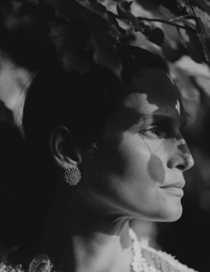 Close-up black and white portrait of an adult woman with short hair and large earrings, standing in shadow with leaves overhead.