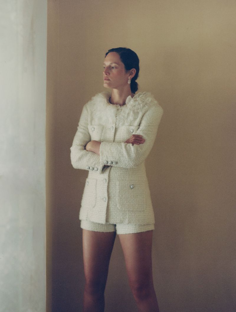 An adult female model in a tweed jacket and shorts suit with her arms crossed, posing in front of a two-toned wall in a photo studio.