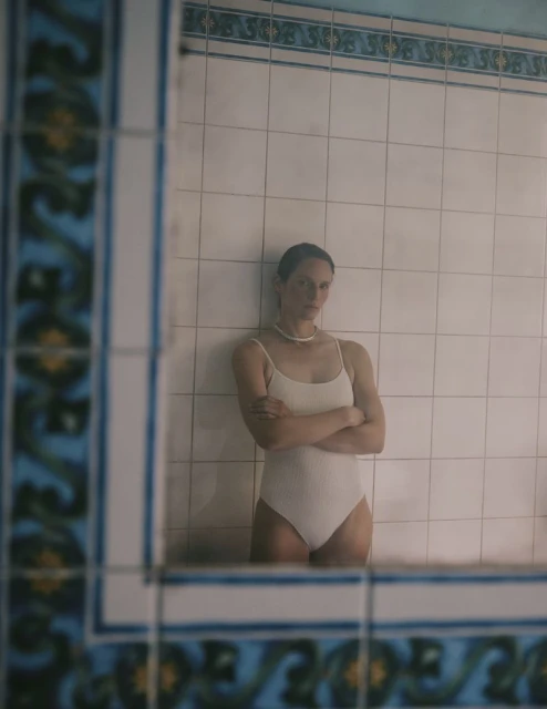 Woman in white bathing suit stands against tiled wall in front of mirror with blue tiled frame in an indoor setting.