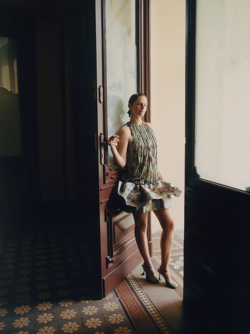 Elegant woman in a patterned dress standing in an open doorway with patterned floor tiles. Fashion shoot, indoor setting, natural light.