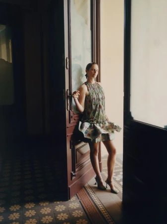 Elegant woman in a patterned dress standing in an open doorway with patterned floor tiles. Fashion shoot, indoor setting, natural light.