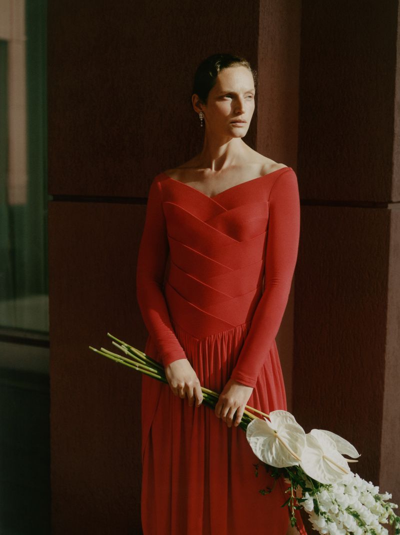 Model Vivien Solari in an off-the-shoulder red long-sleeve dress holding a bouquet of white flowers in directional sunlight.