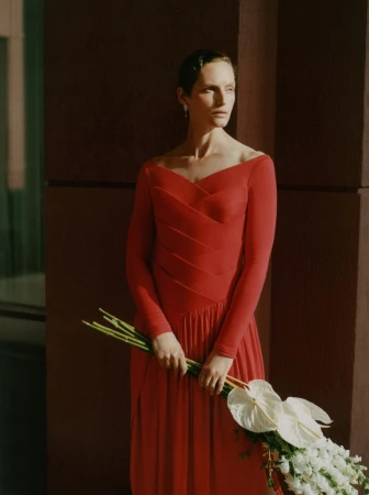 Model Vivien Solari in an off-the-shoulder red long-sleeve dress holding a bouquet of white flowers in directional sunlight.