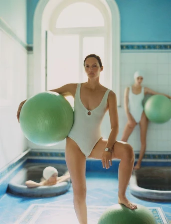 Indoor view of three women in bathing suits and swim caps, exercising with green exercise balls in a blue and white tiled room.