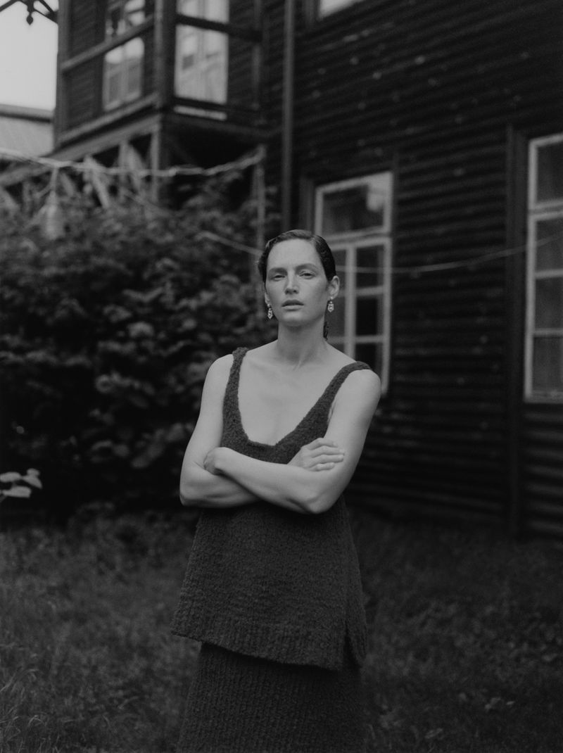 Monochromatic portrait of a woman in a knitted dress with her arms crossed, standing outside with trees and a weathered building behind her.