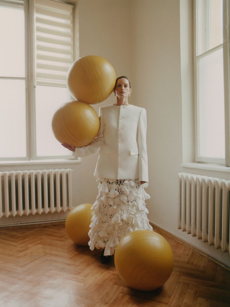 Woman in white jacket and skirt poses with yellow balls in a room with parquet flooring and windows, creating a surreal fashion image.