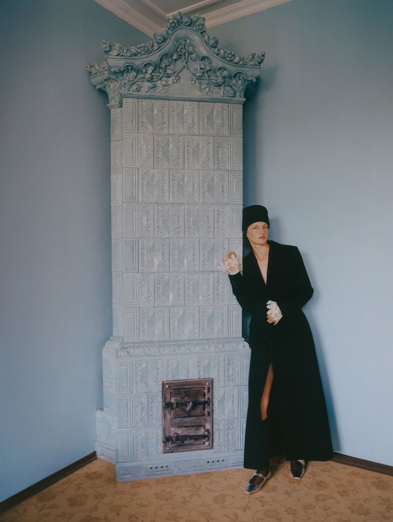 A woman in a black coat and hat stands in a room with pale blue walls, leaning against a large, ornate, gray tiled fireplace.