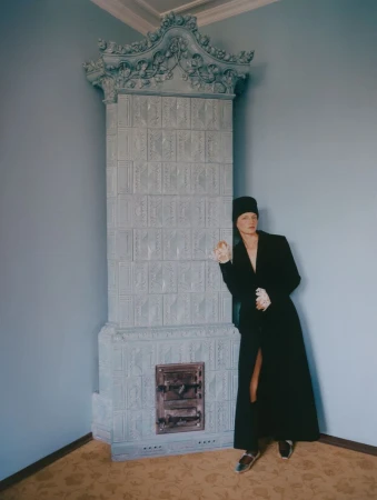A woman in a black coat and hat stands in a room with pale blue walls, leaning against a large, ornate, gray tiled fireplace.