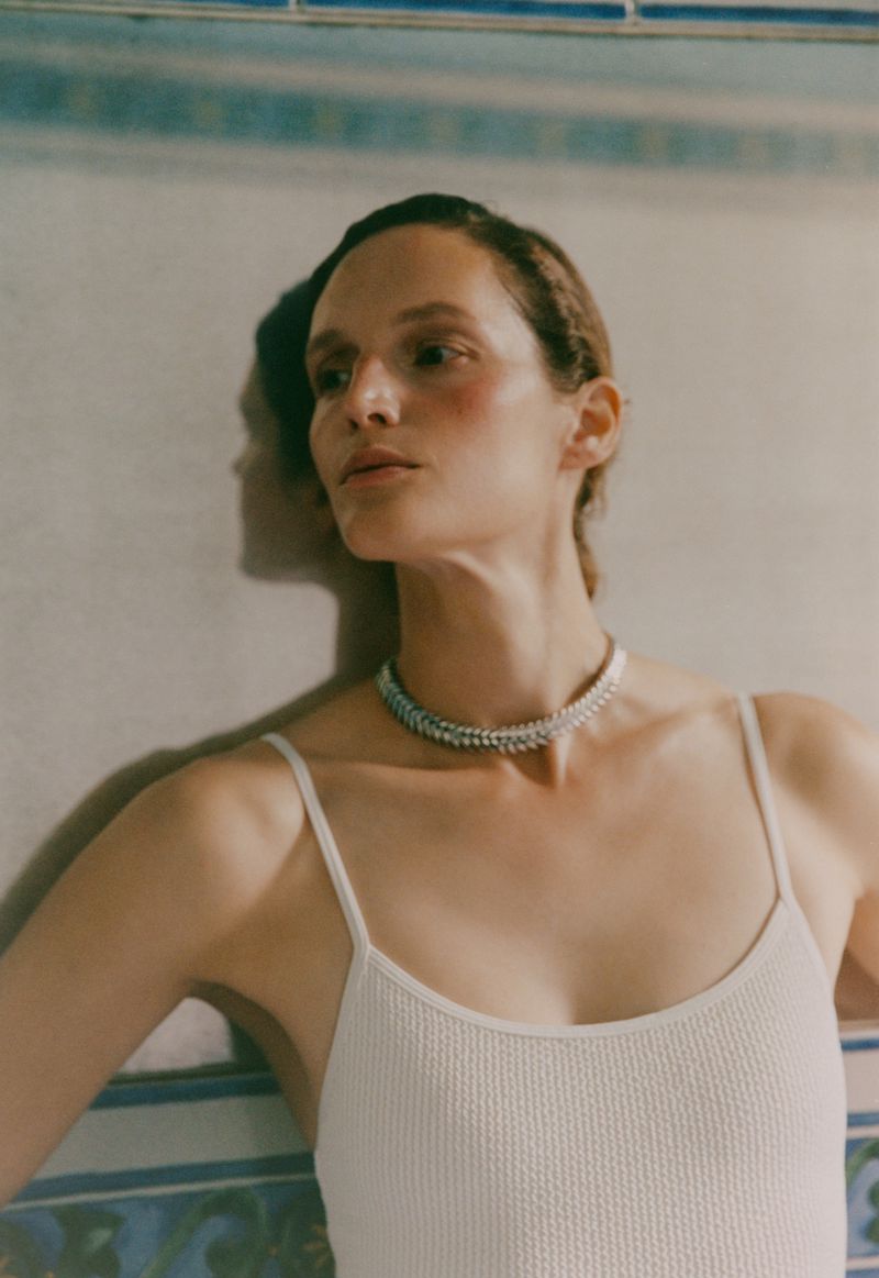 Indoor close-up of a young woman with a neutral expression, wearing a white knit top and statement jewelry, with a tiled wall background.
